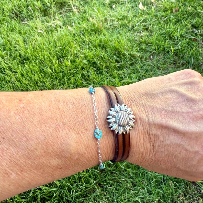 May include: A brown leather wrap bracelet with a silver sunflower charm and a silver chain bracelet with turquoise beads. The bracelets are displayed on a person's wrist against a background of green grass.