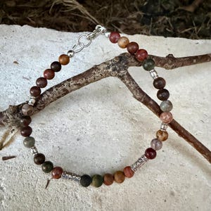 May include: A beaded bracelet featuring round, multi-colored earth-toned stones in shades of brown, green, and red. The bracelet is accented with small silver-toned metal spacers and two cylindrical, textured silver-toned metal beads, finished with a silver-toned lobster claw clasp.