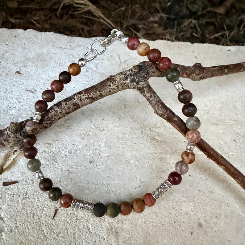 May include: A beaded bracelet featuring round, multi-colored earth-toned stones in shades of brown, green, and red. The bracelet is accented with small silver-toned metal spacers and two cylindrical, textured silver-toned metal beads, finished with a silver-toned lobster claw clasp.