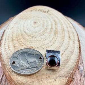 Puede incluir: Un anillo de plata con una piedra preciosa de color rojo oscuro y una moneda de plata con un dise&ntilde;o de b&uacute;falo. El anillo tiene una textura martillada y un engaste de bisel. La moneda es una moneda de cinco centavos de los Estados Unidos.