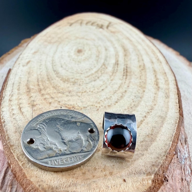 Puede incluir: Un anillo de plata con una piedra preciosa de color rojo oscuro y una moneda de plata con un dise&ntilde;o de b&uacute;falo. El anillo tiene una textura martillada y un engaste de bisel. La moneda es una moneda de cinco centavos de los Estados Unidos.