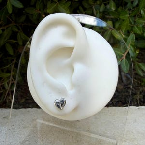May include: A small, silver-toned heart-shaped stud earring with a textured, ridged surface. The earring is displayed on a white, ear-shaped mannequin stand, showcasing the metallic finish and the detailed, vertical line pattern on the heart.