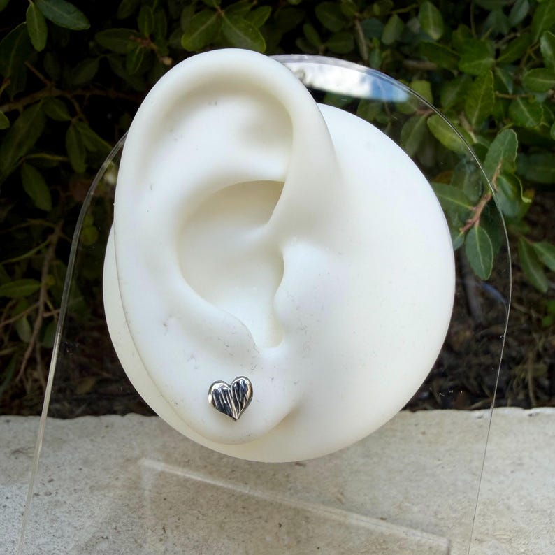 May include: A small, silver-toned heart-shaped stud earring with a textured, ridged surface. The earring is displayed on a white, ear-shaped mannequin stand, showcasing the metallic finish and the detailed, vertical line pattern on the heart.