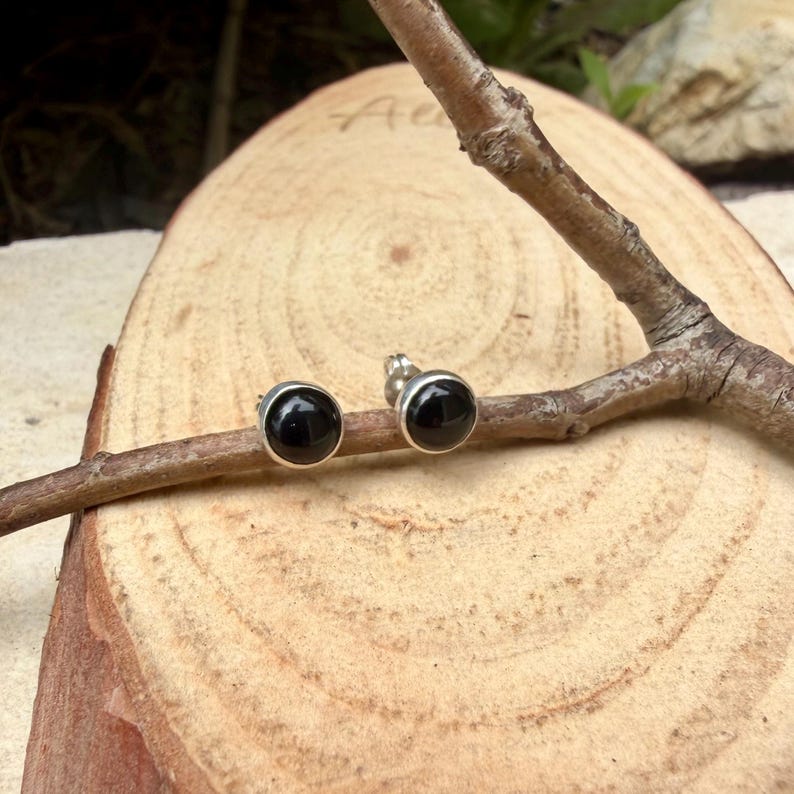 May include: A pair of small, round stud earrings with black, polished stones set in silver-colored metal. The earrings are displayed on a wooden surface with a natural branch.