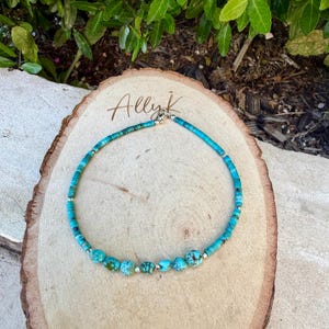 May include: Turquoise beaded necklace with silver accents. The necklace features a mix of small, round turquoise beads and larger, irregularly shaped turquoise stones. The necklace is displayed on a wooden surface with the handwritten text "Allyk".