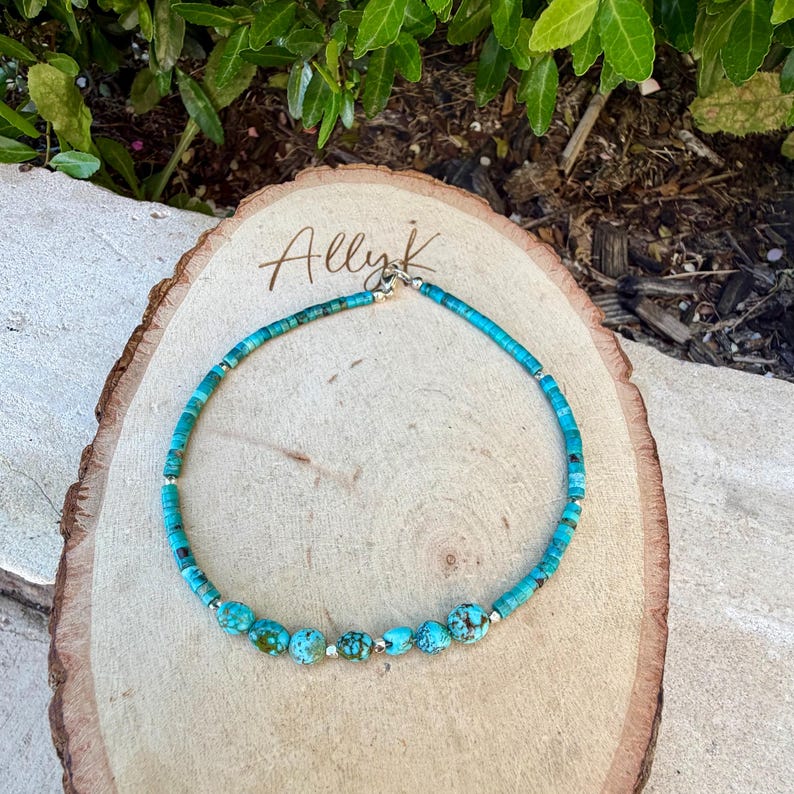 May include: Turquoise beaded necklace with silver accents. The necklace features a mix of small, round turquoise beads and larger, irregularly shaped turquoise stones. The necklace is displayed on a wooden surface with the handwritten text "Allyk".