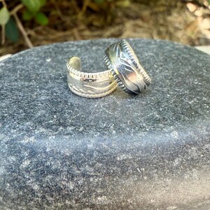May include: Two silver-toned open rings featuring a textured band with a repeating leaf-like pattern and a notched edge. The metallic bands are curved and display a polished finish with darkened recessed areas to highlight the intricate design.