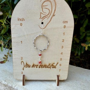 May include: A silver hoop earring with a dangling charm featuring two small red beads and a silver spacer bead. The earring is displayed on a light wood stand with an ear outline, inch and cm markings, and the text "You are beautiful".