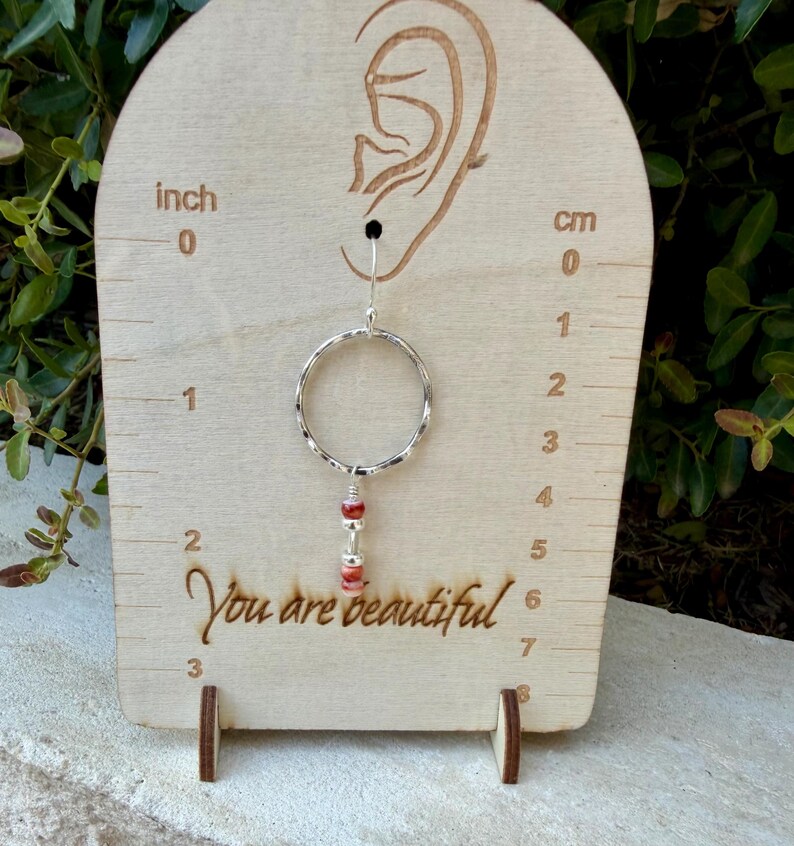May include: A silver hoop earring with a dangling charm featuring two small red beads and a silver spacer bead. The earring is displayed on a light wood stand with an ear outline, inch and cm markings, and the text "You are beautiful".
