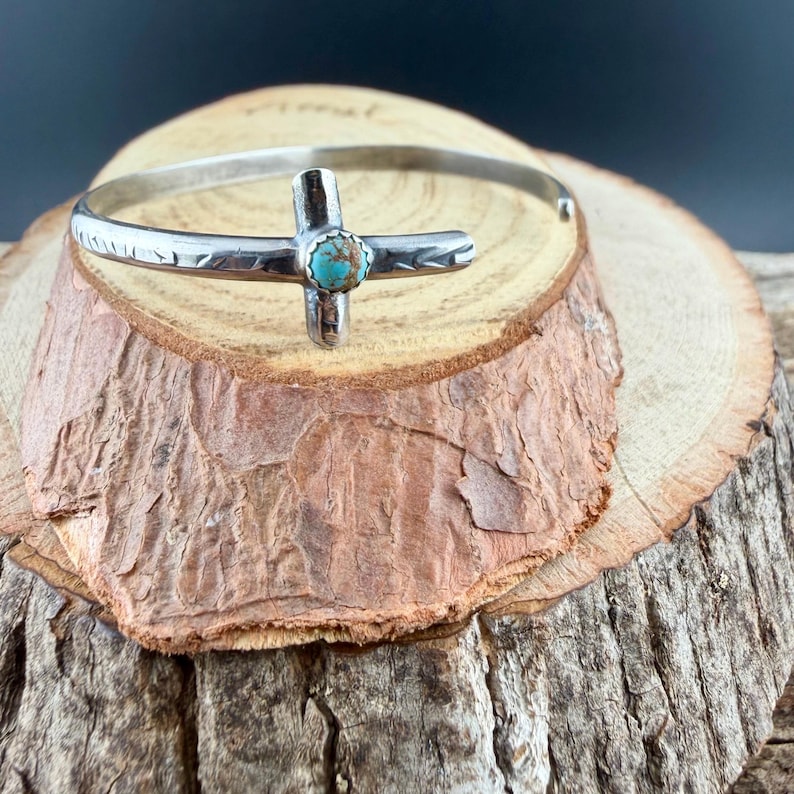 May include: A silver-toned cuff bracelet with a cross-shaped centerpiece. The cross features a turquoise stone at its center. The bracelet is displayed on a wooden surface, showcasing its handcrafted details and Southwestern style.