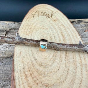 May include: A gold-colored ring with a turquoise stone is displayed on a wooden surface. The ring is rectangular with a rounded turquoise stone. The wooden surface has the handwritten text "Allyk".
