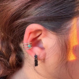 May include: Close-up of an ear adorned with two pieces of jewelry. A silver ear cuff with three bands and a turquoise stone sits on the upper ear. A small black hoop earring hangs from the earlobe.
