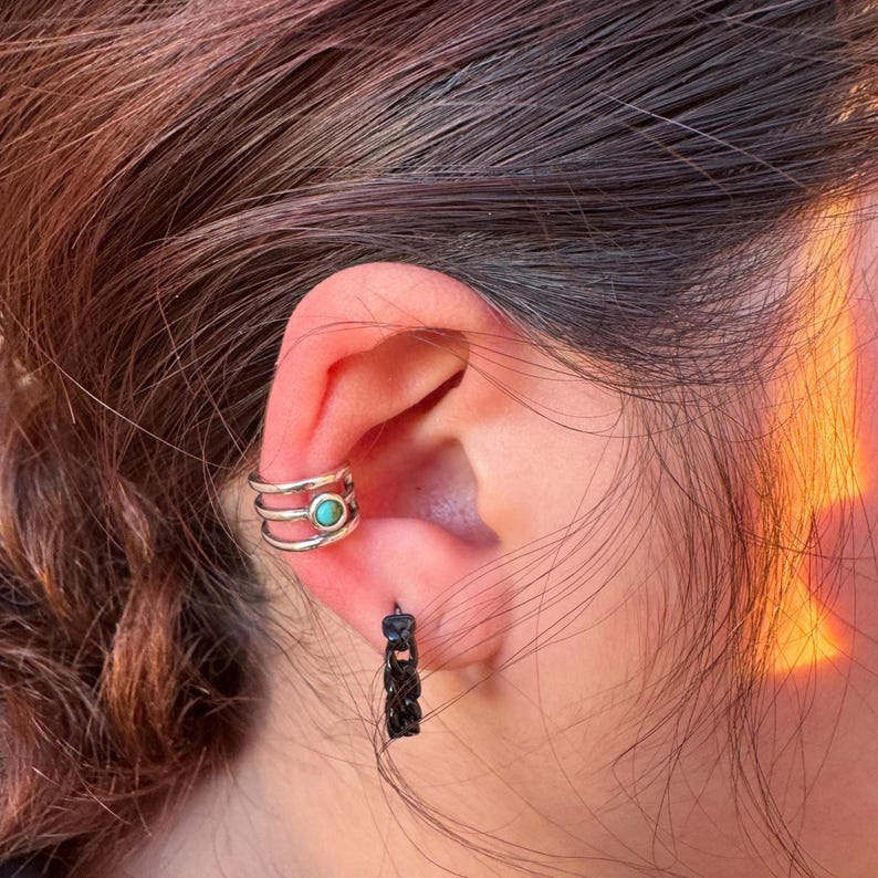 May include: Close-up of an ear adorned with two pieces of jewelry. A silver ear cuff with three bands and a turquoise stone sits on the upper ear. A small black hoop earring hangs from the earlobe.