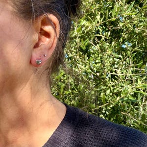 May include: A small, polished silver-toned heart-shaped stud earring worn in the earlobe. The earring features a smooth, reflective metallic surface and a simple, compact design that sits flush against the skin of the ear.