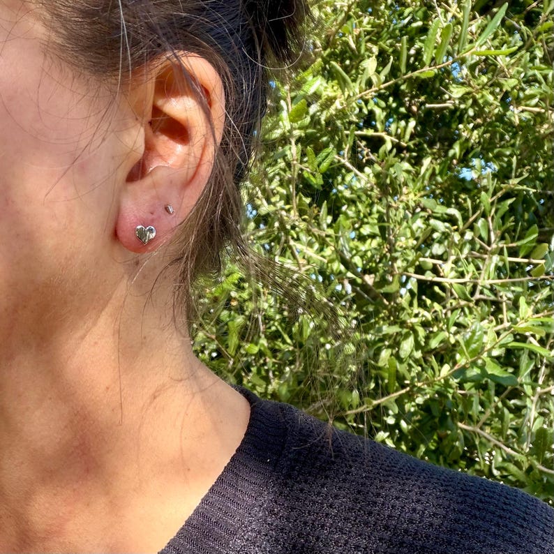May include: A small, polished silver-toned heart-shaped stud earring worn in the earlobe. The earring features a smooth, reflective metallic surface and a simple, compact design that sits flush against the skin of the ear.