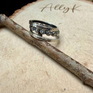 May include: A stackable silver ring set with three bands. The top band has a floral design, the middle band has a beaded design, and the bottom band has an etched pattern. The rings are displayed on a wooden surface.