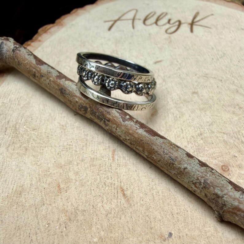 May include: A stackable silver ring set with three bands. The top band has a floral design, the middle band has a beaded design, and the bottom band has an etched pattern. The rings are displayed on a wooden surface.