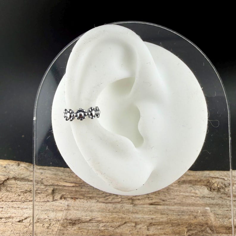 Puede incluir: Un ear cuff plateado con un dise&ntilde;o floral. La joya se muestra en un modelo de oreja blanco, resaltando sus detalles. El fondo combina texturas negras y de madera, acentuando las caracter&iacute;sticas del ear cuff.