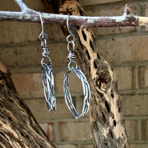 Op de afbeelding: Een paar zilverkleurige hangende oorbellen met een gevlochten metalen ontwerp. Elke oorbel bestaat uit een cirkelvormig ringelement dat aan een eenvoudige haak hangt, met een gewikkeld metalen detail dat de haak met de ring verbindt.
