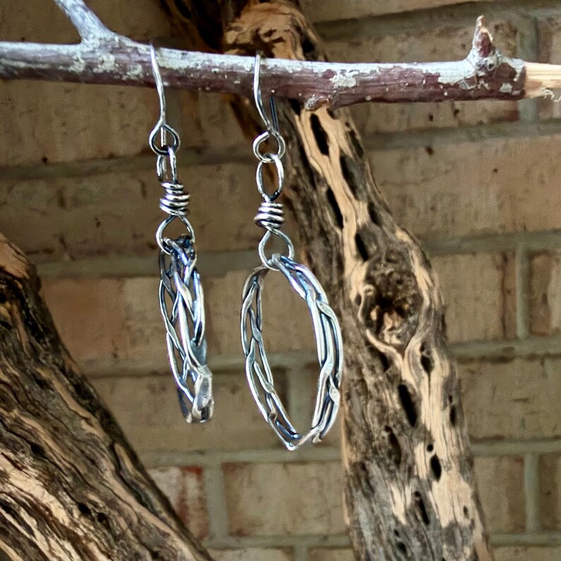 Op de afbeelding: Een paar zilverkleurige hangende oorbellen met een gevlochten metalen ontwerp. Elke oorbel bestaat uit een cirkelvormig ringelement dat aan een eenvoudige haak hangt, met een gewikkeld metalen detail dat de haak met de ring verbindt.