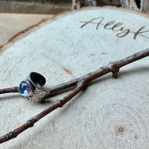 May include: A silver ring with a large, iridescent, round gemstone. The ring has an engraved design on the band and is displayed on a wooden slice with the name "Allyk" etched into the wood. A small twig rests across the wood.