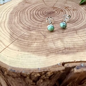 Pu&ograve; includere: Orecchini a lobo in argento con design floreale e pietre rotonde verde chiaro. Gli orecchini sono esposti su una superficie di legno con anelli di crescita visibili.