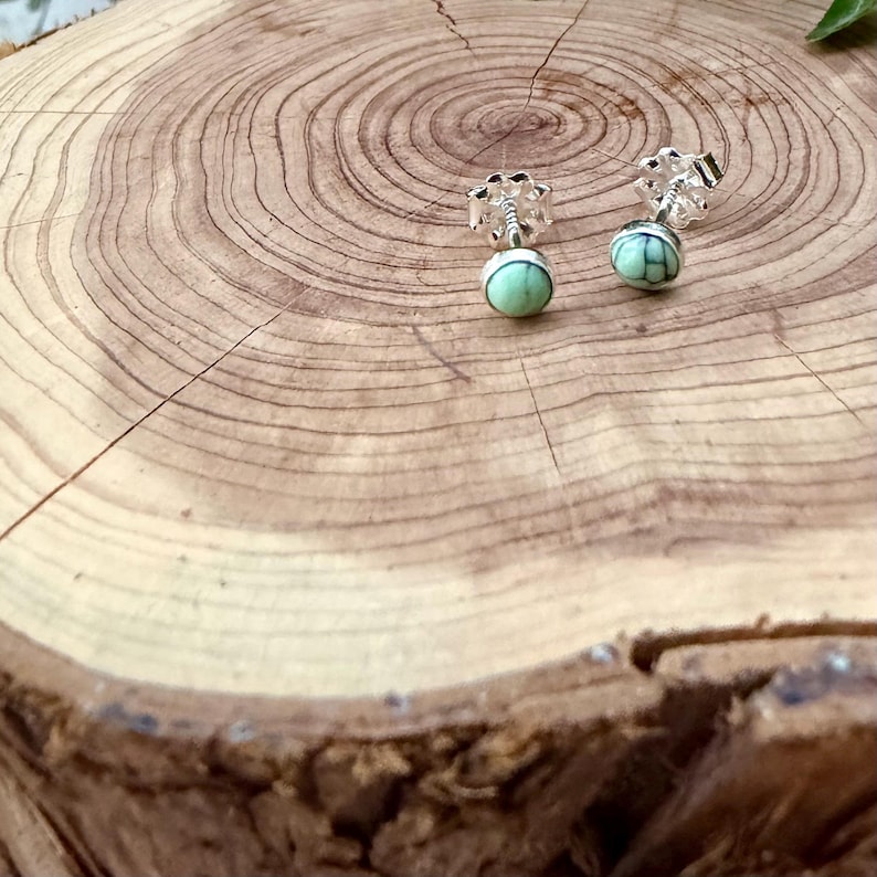 Pu&ograve; includere: Orecchini a lobo in argento con design floreale e pietre rotonde verde chiaro. Gli orecchini sono esposti su una superficie di legno con anelli di crescita visibili.