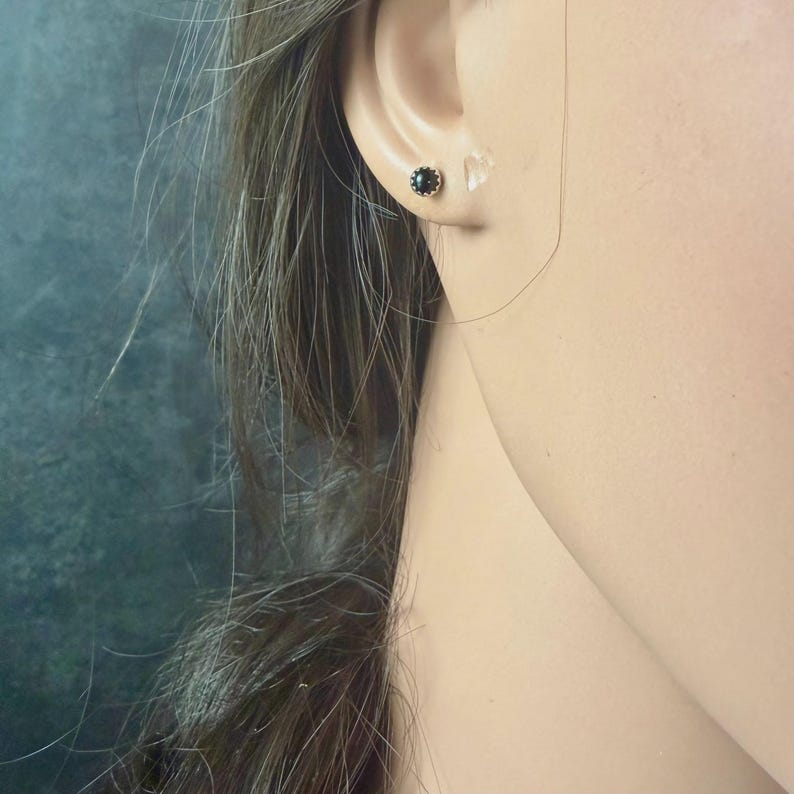 May include: Close-up of a person's ear with two small stud earrings. One earring features a dark, possibly black, stone set in a gold-colored metal. The other earring is a clear, possibly crystal, stud. The background is a muted teal color.