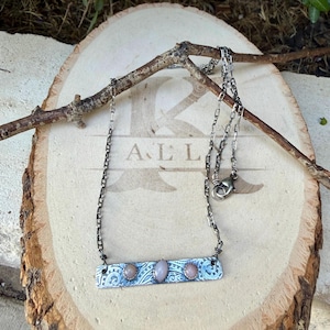 May include: A silver-toned metal bar necklace featuring three small, round, pale pink cabochon stones set in a row. The rectangular bar has an intricate, etched swirl pattern and is suspended from a dark, oxidized silver-toned chain.