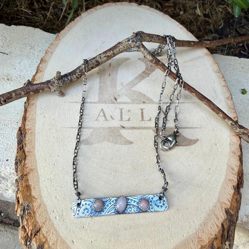 May include: A silver-toned metal bar necklace featuring three small, round, pale pink cabochon stones set in a row. The rectangular bar has an intricate, etched swirl pattern and is suspended from a dark, oxidized silver-toned chain.