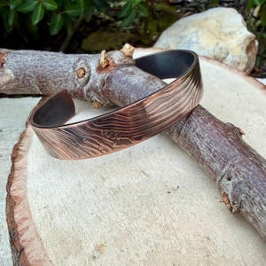 May include: A copper-colored cuff bracelet with a textured, wave-like pattern. The inside of the bracelet is dark. The bracelet is displayed on a piece of wood and a tree branch.