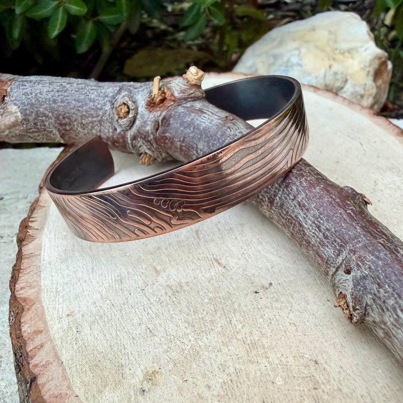 May include: A copper-colored cuff bracelet with a textured, wave-like pattern. The inside of the bracelet is dark. The bracelet is displayed on a piece of wood and a tree branch.
