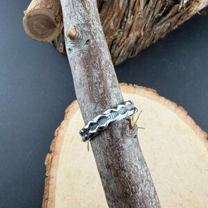 May include: A silver-toned metal ring featuring a continuous, repeating wave-like pattern along the band. The ring has a textured, oxidised finish within the recessed areas of the design, creating a contrast against the polished, raised edges.