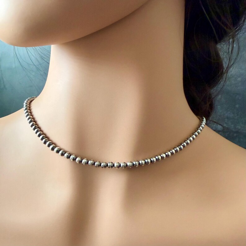 May include: A silver beaded necklace. The necklace is composed of small, round, silver beads strung together to form a choker style necklace. The beads have a slightly textured surface and reflect light, creating a subtle shine. The necklace is displayed on a person's neck.