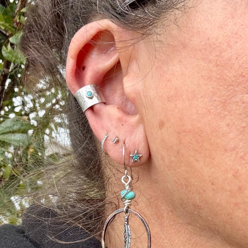 K&ouml;nnte beinhalten: Eine Sammlung von silbernen Ohrringen und einem Ohr-Cuff. Der Ohr-Cuff hat einen T&uuml;rkisstein. Die Ohrringe umfassen einen kleinen sternf&ouml;rmigen Stecker mit einem T&uuml;rkisstein, einen kleinen Stecker, einen Creolen-Ohrring und einen gro&szlig;en Creolen-Ohrring mit einem Federanh&auml;nger.