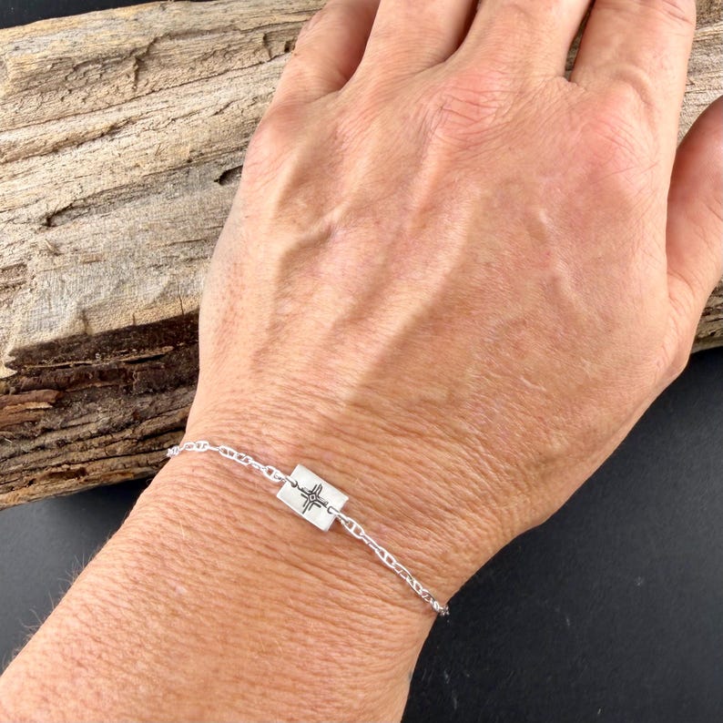 May include: A silver bracelet featuring a rectangular charm with a cross-like design. The bracelet is made of delicate chain links and is displayed on a person's wrist.