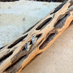 May include: Two silver-toned arrowhead stud earrings with textured surfaces. The earrings are displayed on a piece of light brown wood with dark brown patterns and holes. The earrings are small and detailed.
