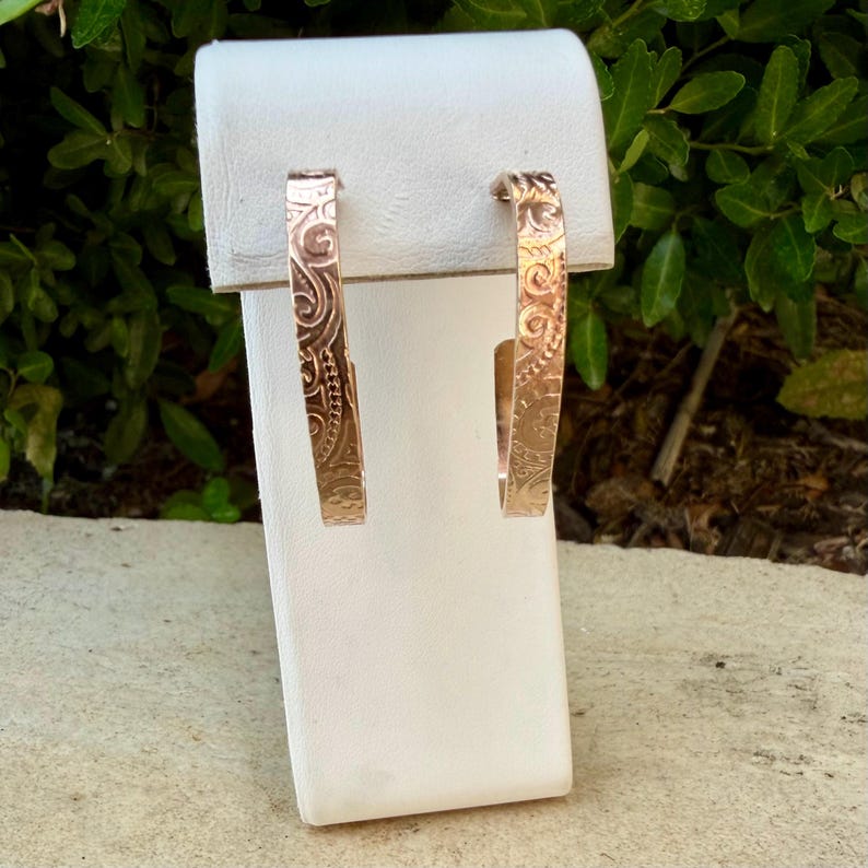 May include: A pair of rose gold hoop earrings with intricate engraved floral and swirl patterns. The earrings are displayed on a white stand against a backdrop of green foliage. The earrings have a rectangular shape.