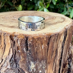 May include: A silver band ring featuring a hammered, textured finish and a prominent black-filled cross symbol engraved on the front surface. The metal has a polished sheen, and the design is consistent around the circumference of the ring.