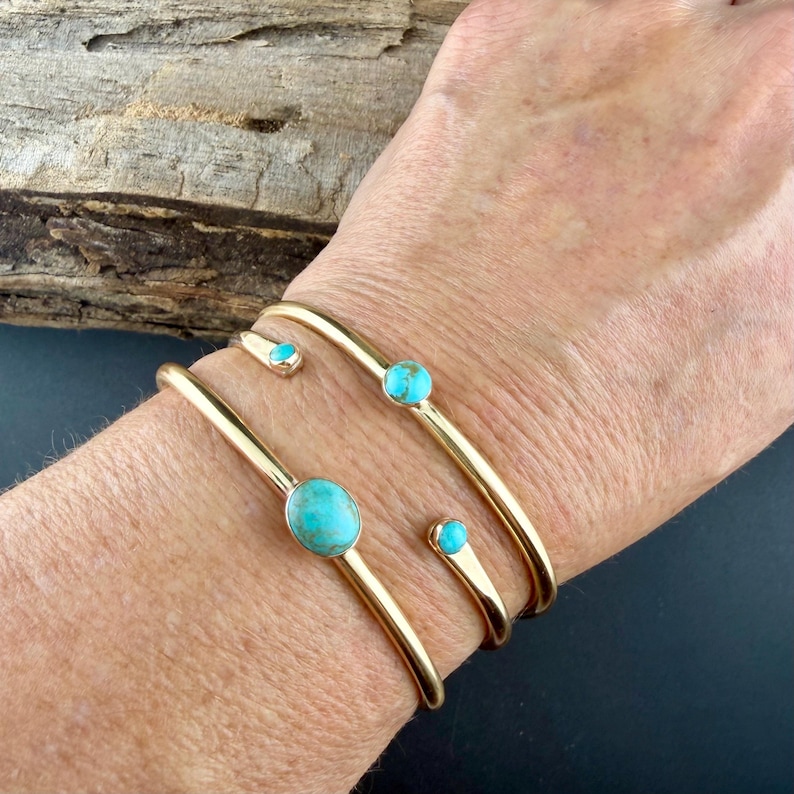 Op de afbeelding: Goudkleurige metalen armbanden met turkooiskleurige stenen. De armbanden zijn in manchetstijl, met &eacute;&eacute;n met een grote ovale steen en andere met kleinere ronde stenen. De armbanden worden om de pols gedragen.