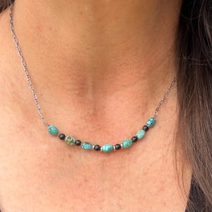 May include: A silver chain necklace featuring turquoise and black beads. The necklace has a delicate chain and is adorned with oval-shaped turquoise beads and small, round black beads. The necklace is designed to be worn around the neck.
