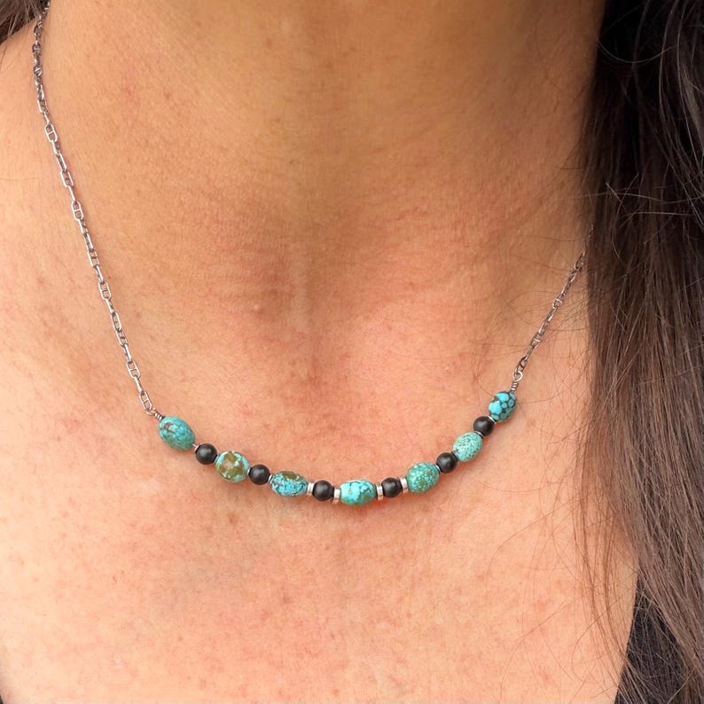 May include: A silver chain necklace featuring turquoise and black beads. The necklace has a delicate chain and is adorned with oval-shaped turquoise beads and small, round black beads. The necklace is designed to be worn around the neck.