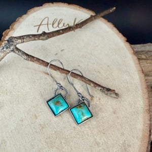 May include: A pair of silver-toned earrings with square turquoise stones. The stones are set in a silver frame and have a mottled appearance. The earrings hang from a twisted silver loop and a simple hook.