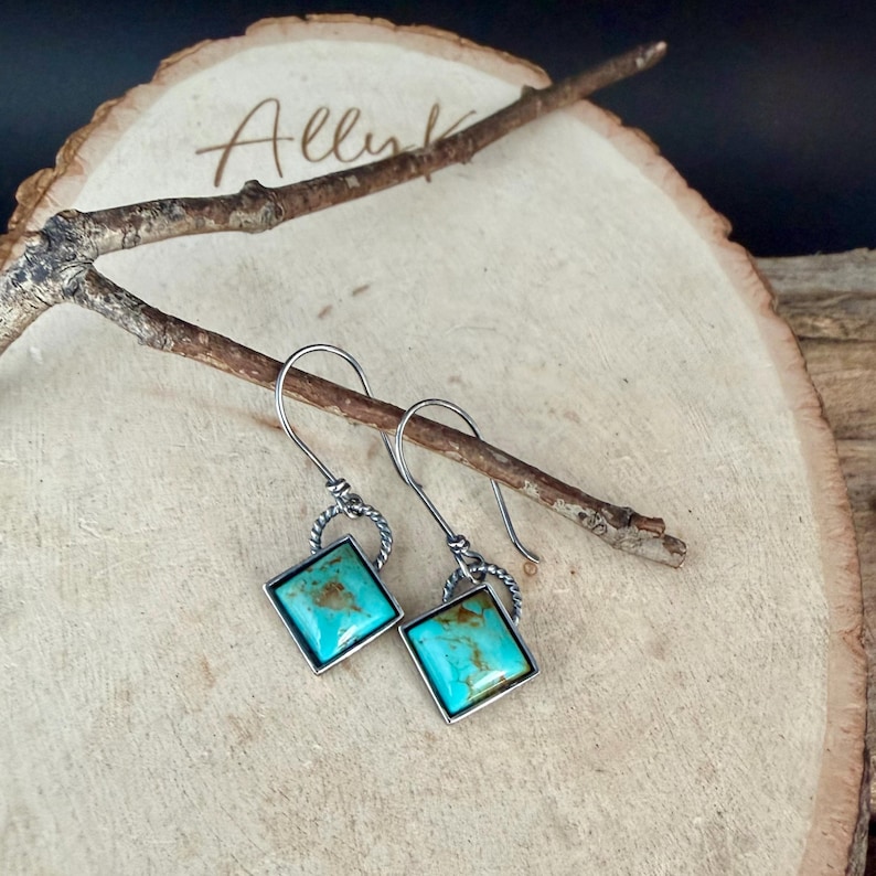 May include: A pair of silver-toned earrings with square turquoise stones. The stones are set in a silver frame and have a mottled appearance. The earrings hang from a twisted silver loop and a simple hook.