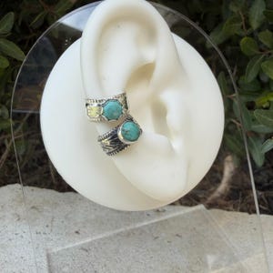 May include: Two silver ear cuffs featuring round turquoise stones set in metal bands with textured, etched detailing. The cuffs are displayed on a white ear-shaped model, showcasing the contrast between the bright blue-green stones and the dark oxidized silver finish.