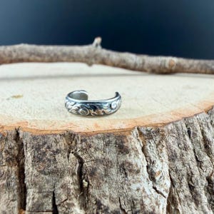 May include: A silver-colored toe ring with an open design. The ring features a detailed floral pattern around the band. The ring is displayed on a rustic wooden surface, showcasing its intricate design and craftsmanship.