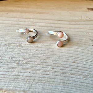 May include: Two open-style rings crafted from silver-toned metal, each featuring two small, round, pale pink cabochon stones set at the ends of the band. The rings have a smooth, polished finish and a simple, adjustable open-ended design.