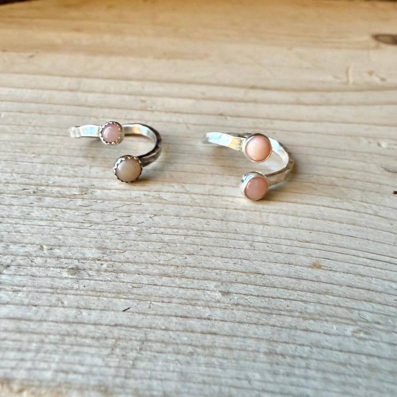 May include: Two open-style rings crafted from silver-toned metal, each featuring two small, round, pale pink cabochon stones set at the ends of the band. The rings have a smooth, polished finish and a simple, adjustable open-ended design.