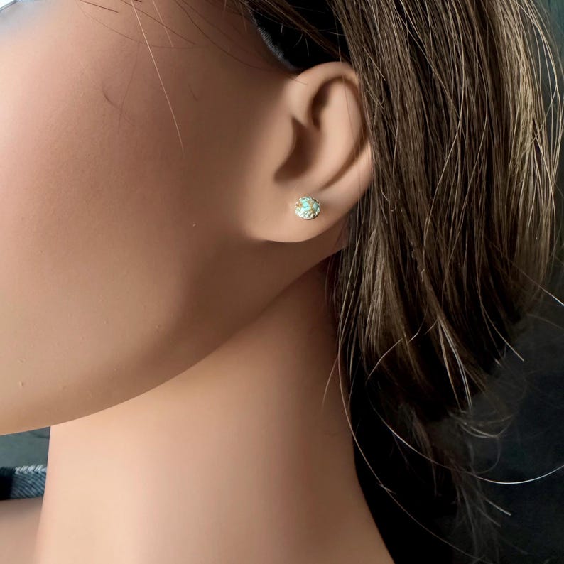 May include: A close-up of a small, round stud earring. The earring features a central light blue stone surrounded by a gold-colored metal setting. The earring is on a person's ear.