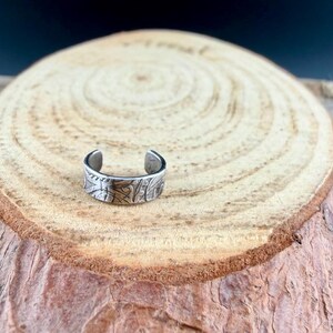 May include: A silver-colored, open-ended ring with an engraved floral pattern. The ring is displayed on a wooden surface with a natural, light brown grain. The ring's design features detailed, textured carvings.
